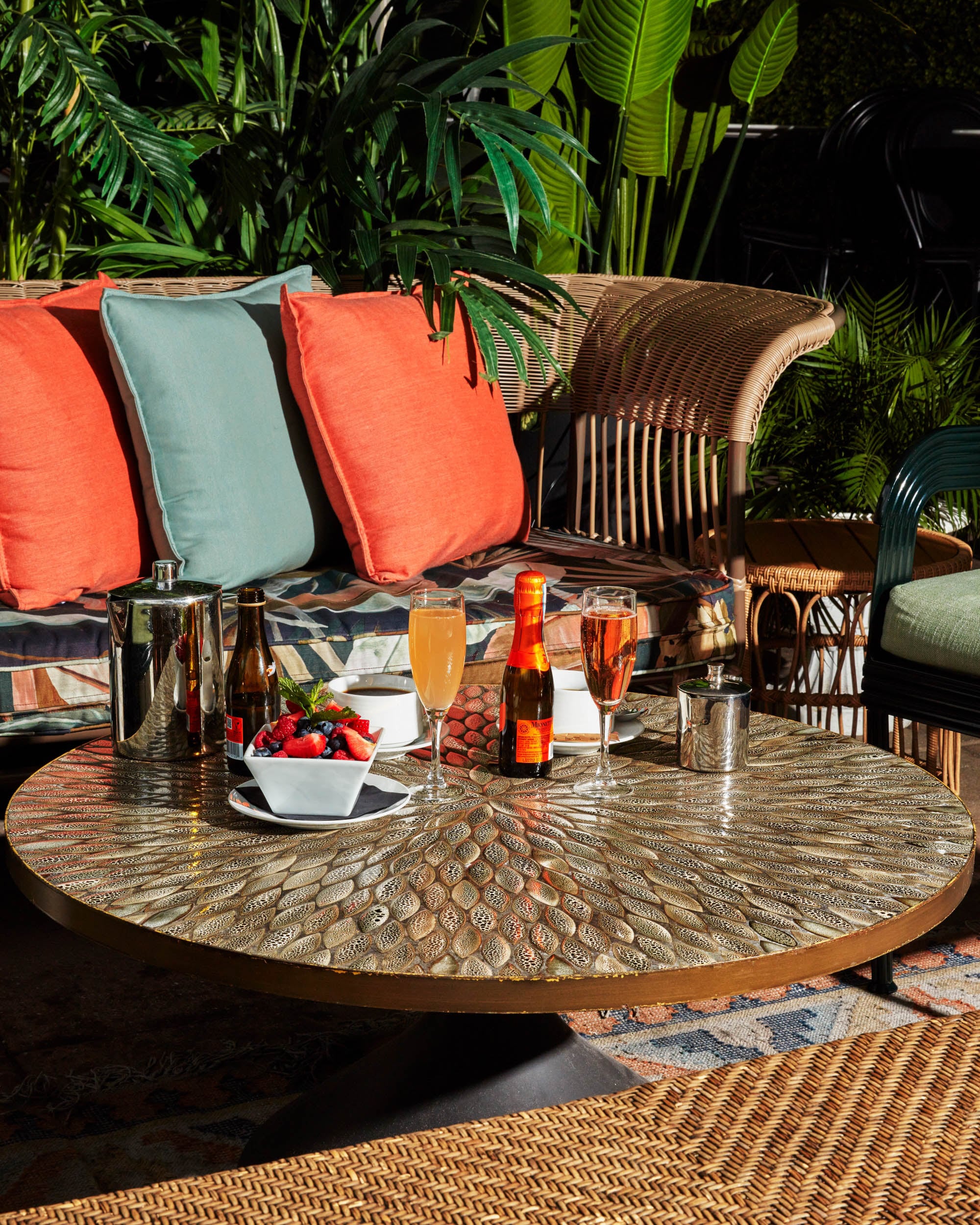 A stylish outdoor dining setup with a round table, wicker furniture, and a selection of drinks and snacks.