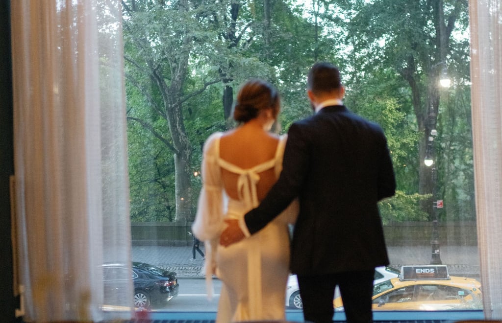 A couple looks out a window at a street with trees and a yellow taxi.