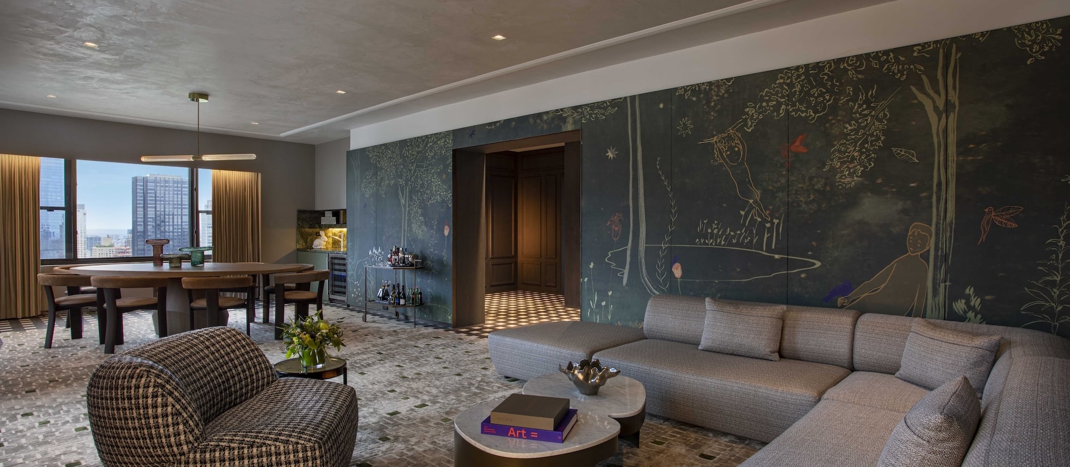 A luxurious living area with a gray sectional sofa, a coffee table, a dining table, and a walled mural, overlooking the city.