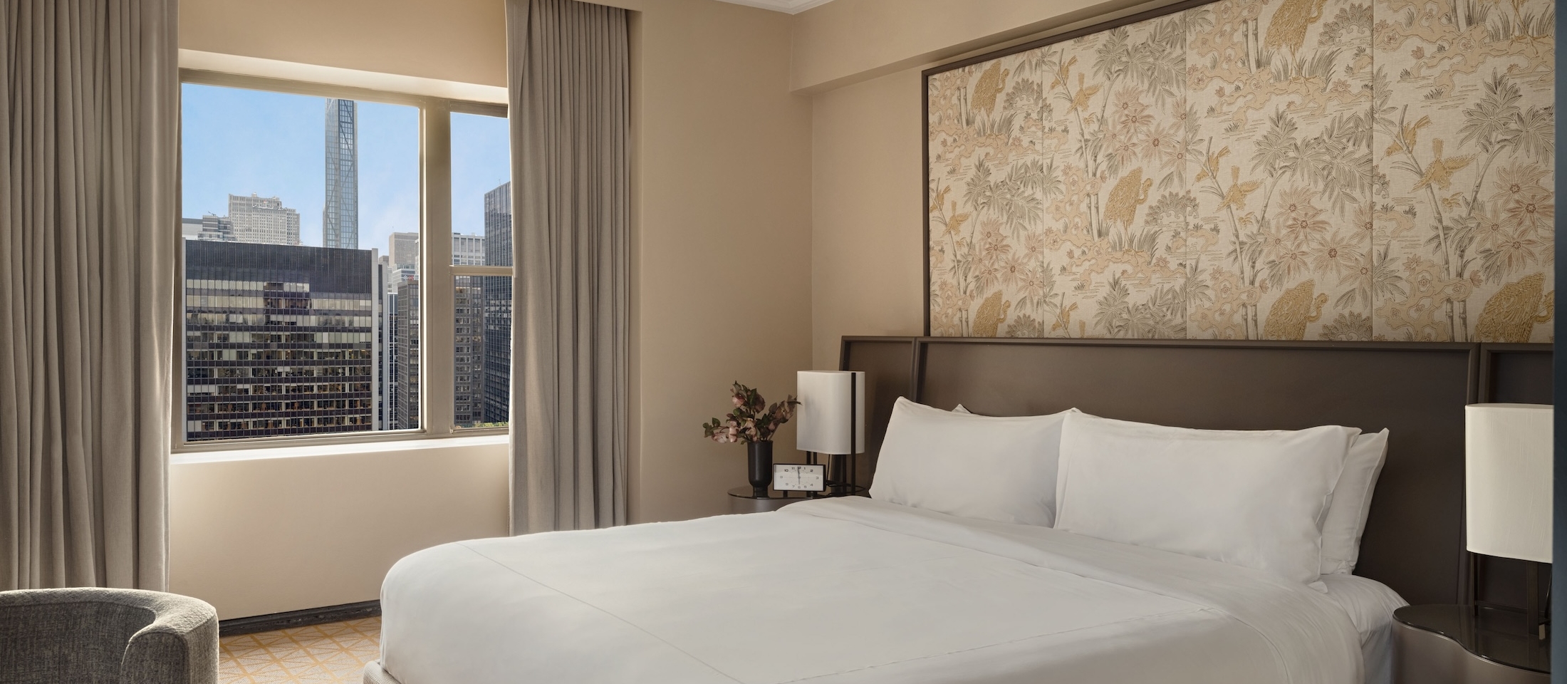 A hotel room with a large bed, a window with a city view, and a floral art piece on the wall.
