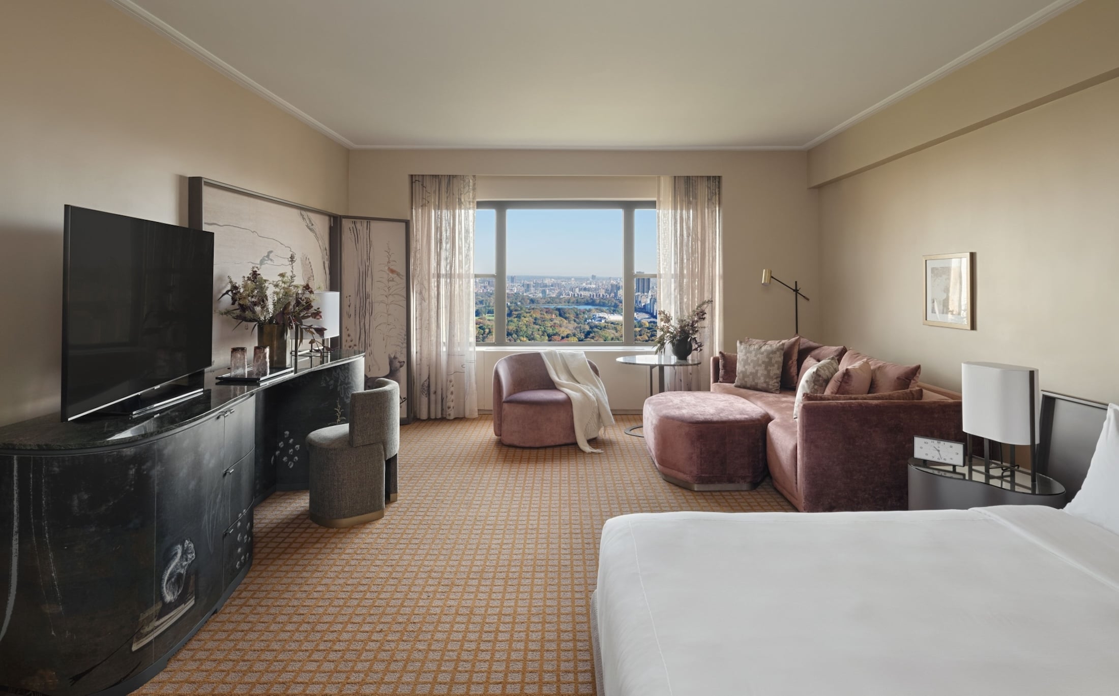 A luxurious hotel suite with a large window offering a city view, a seating area with a sofa and armchair, and a bed.