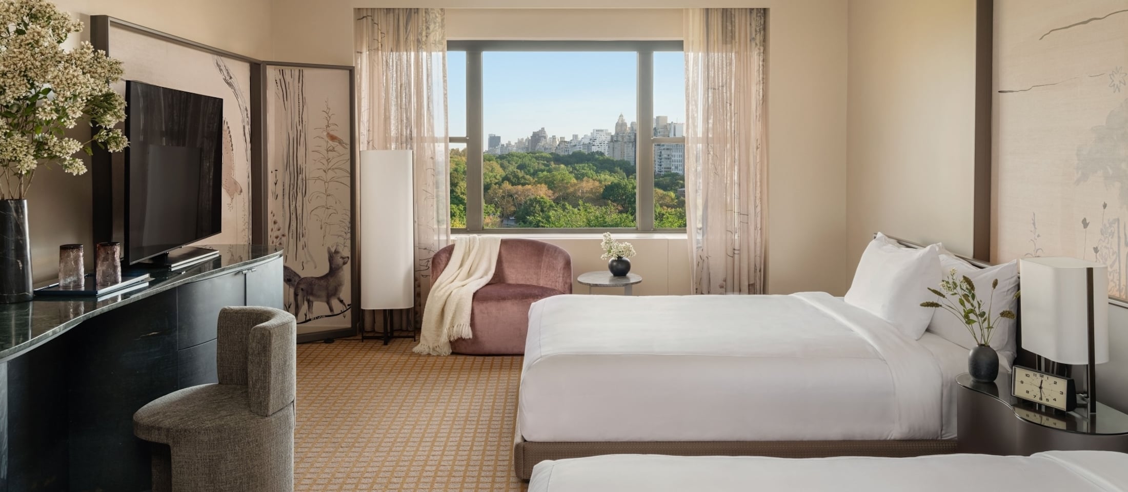 A hotel room with two queen beds, a TV, dresser, and armchair with a city view out the window.