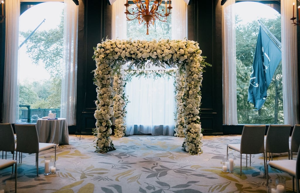 A floral arrangement in a room with a chandelier, windows, chairs, and a table.