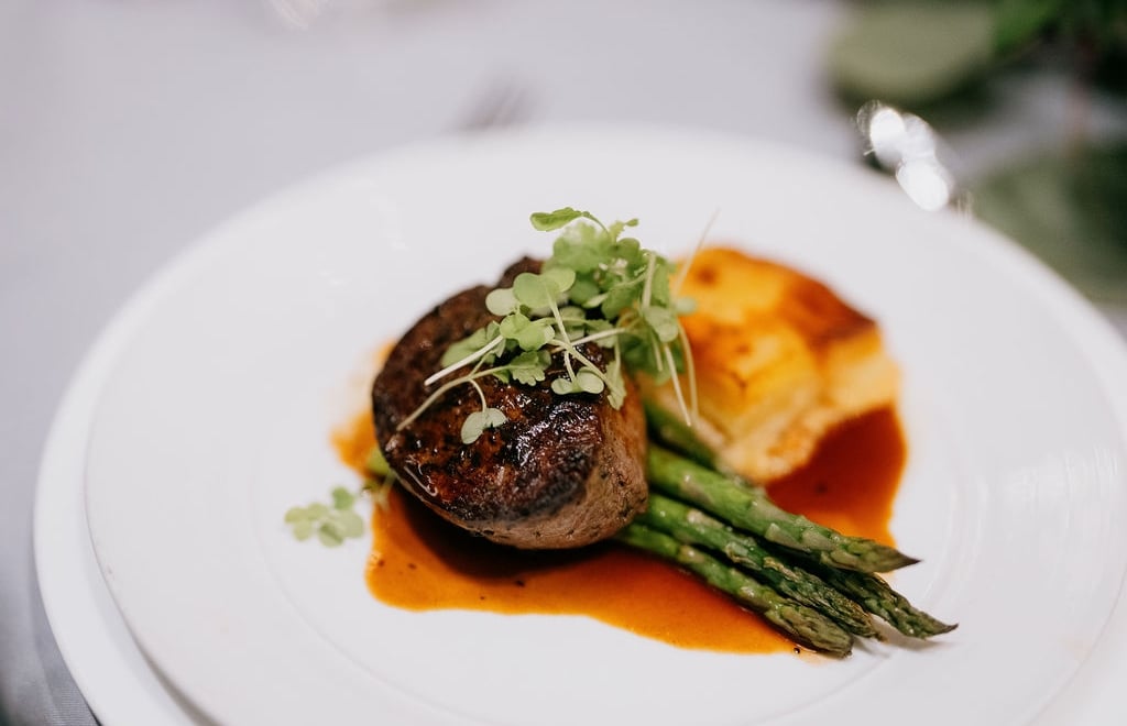 A plated meal consisting of a piece of meat, asparagus, and a side dish, garnished with microgreens and covered in sauce.