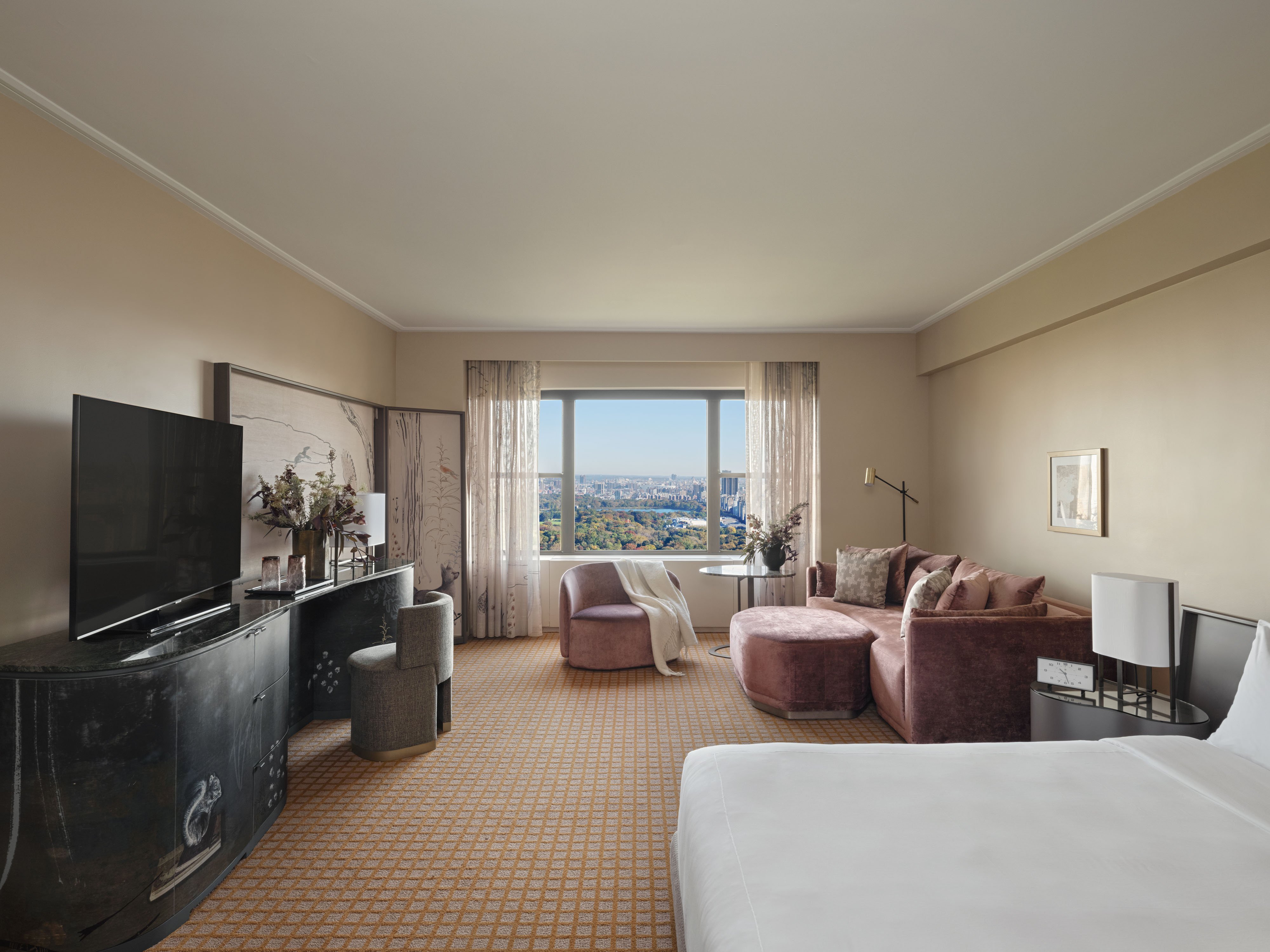 A wide-angle view of a hotel room with a city view, featuring a large bed, sofa, armchair, and window.