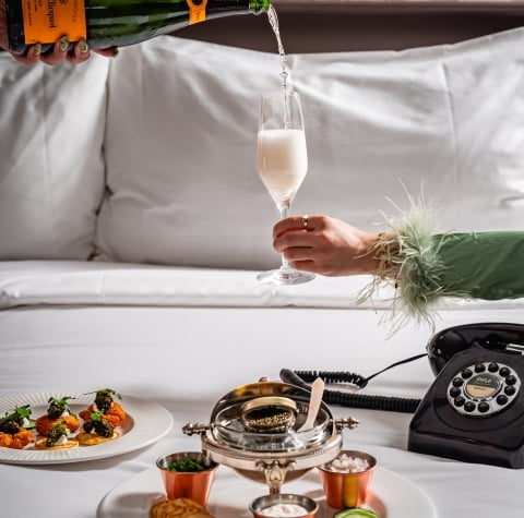 A bedside table with a plate of food, a champagne bottle being poured into a glass, and a black phone.