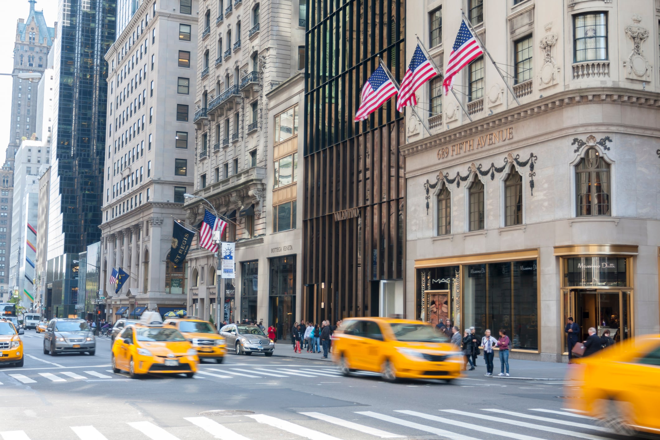 A wide exterior view of a building in New York City with American flags and yellow taxis passing by.